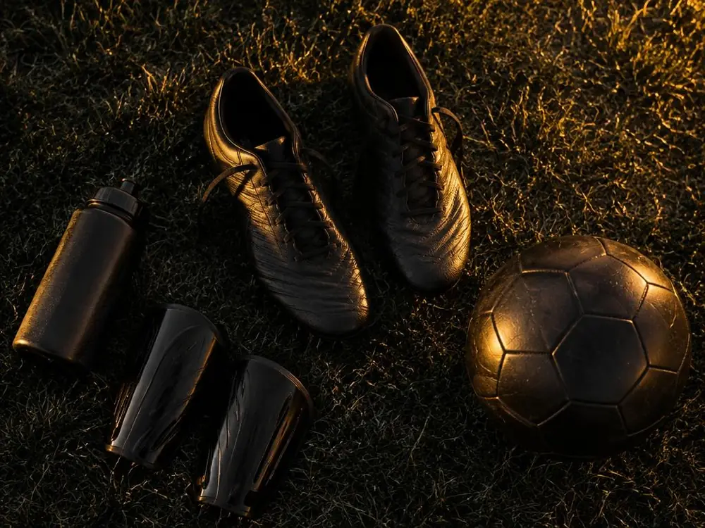 Crampons, protège-tibias, gourde et ballon posés sur une pelouse sombre sous une lumière dorée